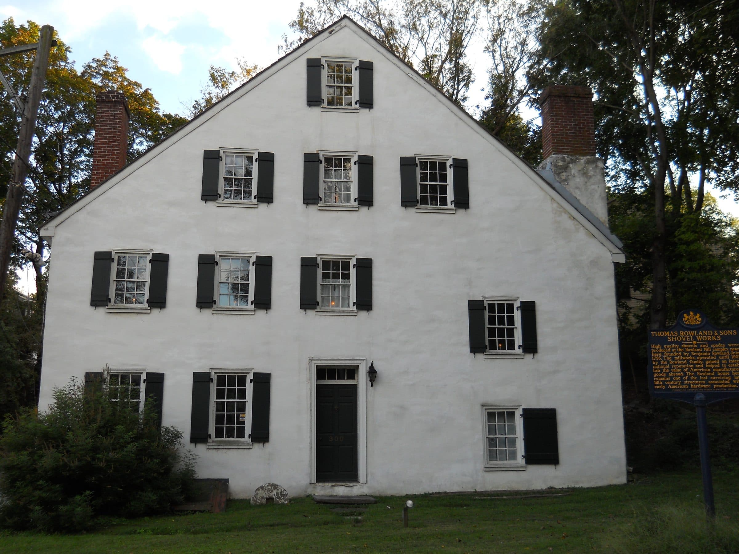 The Rowland House in Cheltenham, Pennsylvania — a tall whitewashed eighteenth-century house with black shutters and two brick chimneys, beside the state marker for the Thomas Rowland & Sons shovel works.