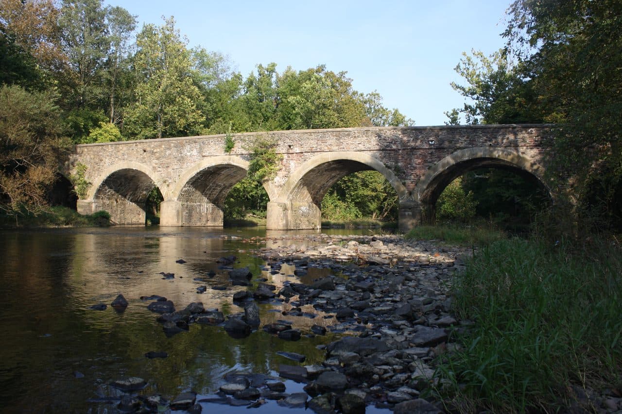 The three-arch stone bridge at Bridge Valley near Furlong, Pennsylvania, crossing a shallow rocky creek in summer.