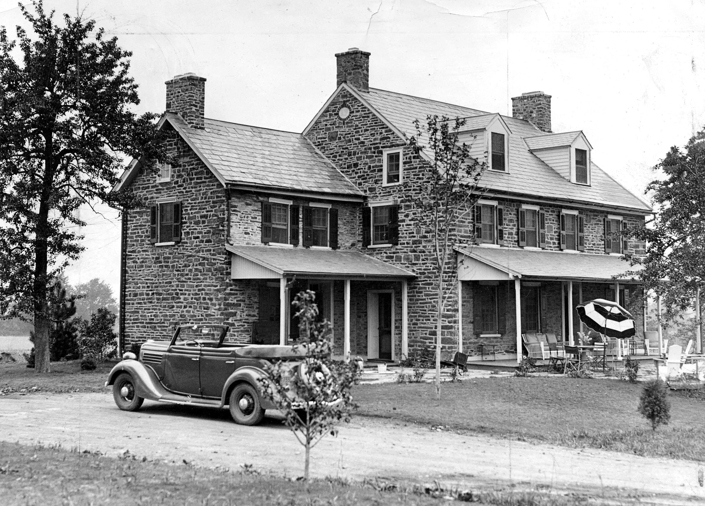 A stone farmhouse at Dresher, Pennsylvania, in an early photograph — a long fieldstone house with three chimneys, shuttered windows and a shingled porch, a period convertible standing on the drive.