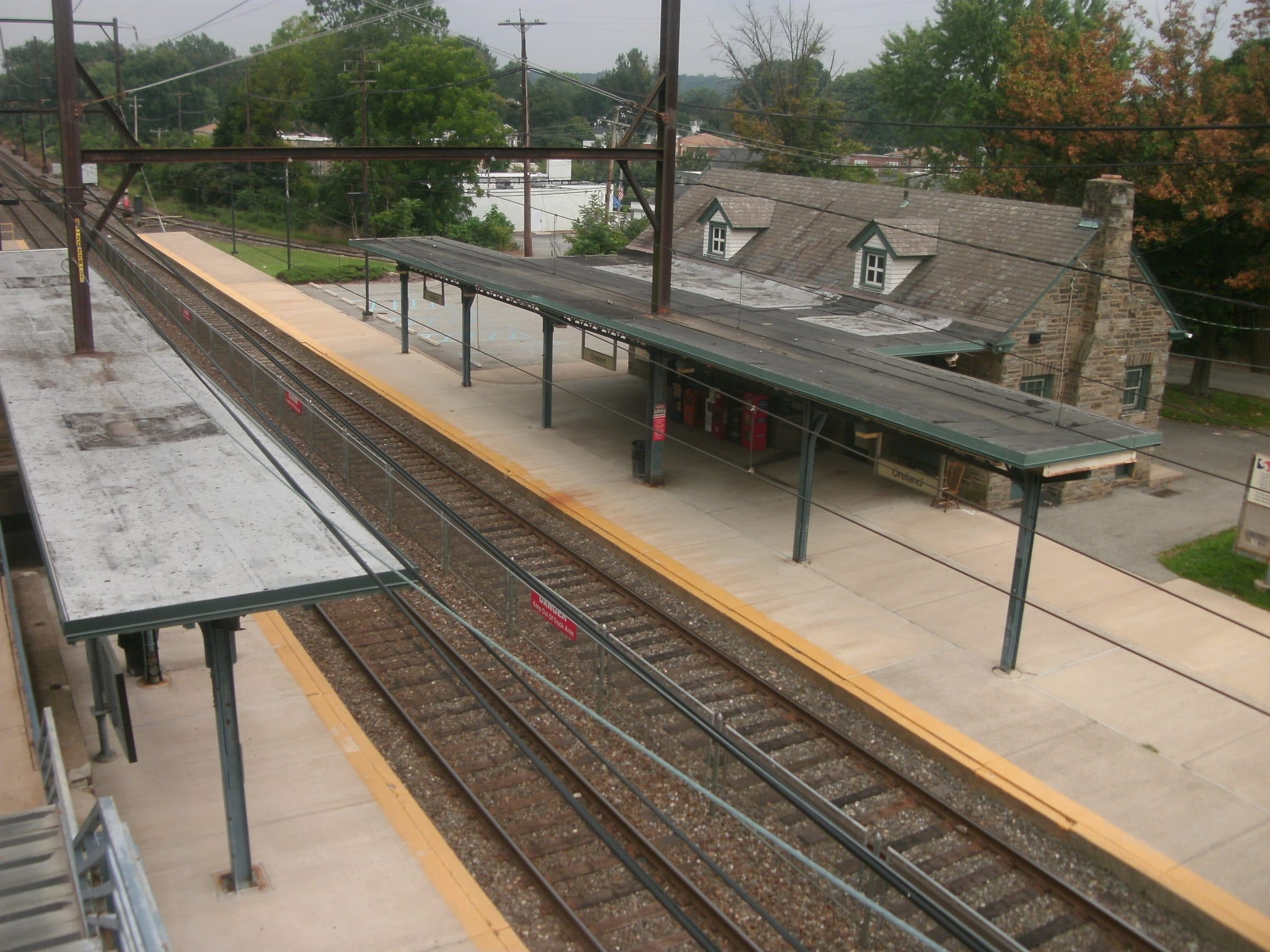 Oreland station in Pennsylvania seen from the footbridge — a stone station building with dormers beside the platform canopy and the tracks of the Lansdale/Doylestown line.