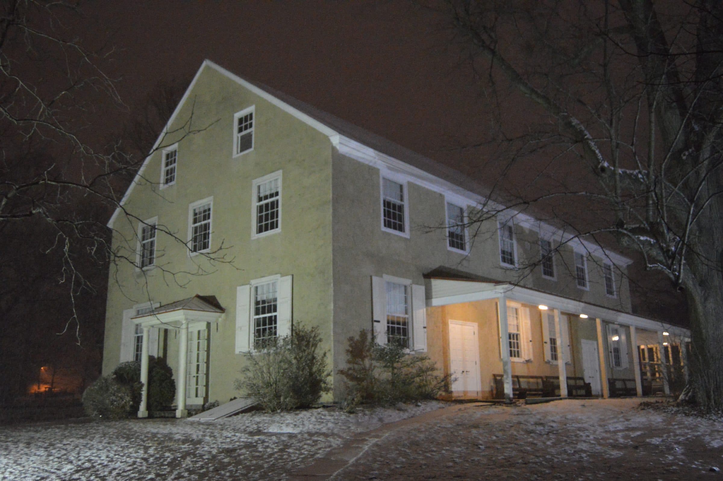 The Friends meeting house in Lower Gwynedd Township, Pennsylvania, lit at night after snow — a plain stuccoed building with a small columned porch and benches under a covered walk.