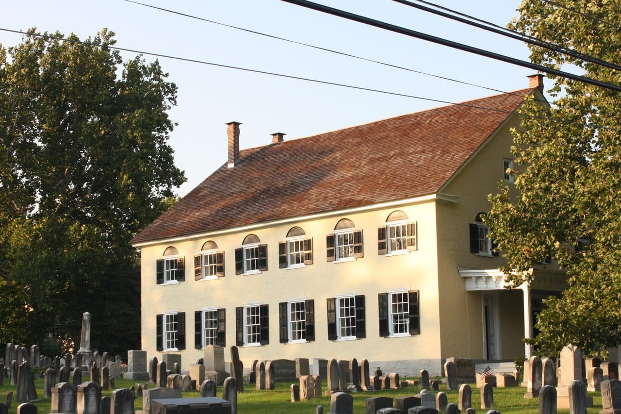 The historic Southampton Baptist Church in Southampton, Pennsylvania, a yellow stuccoed meetinghouse with a red shingled roof and rows of headstones in front.